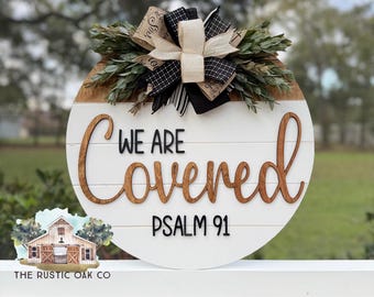 May include: A round wooden sign with the words "WE ARE Covered" and "PSALM 91" in a mix of brown and black lettering. The sign is decorated with a bow and greenery. The sign is white with a wooden top.