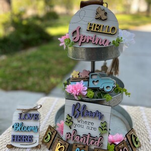 May include: A two-tiered metal tray with a wooden sign that says "Hello Spring" in pink letters. The tray is decorated with pink flowers, a wooden truck, and a wooden banner that says "Spring". There are also two smaller wooden signs that say "Love Blooms Here" and "Bloom Where You Are Planted".
