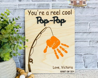 May include: A wooden sign with the text "You're a reel cool Pap-Pap" and a handprint in orange paint. The sign also includes the text "Love, Victoria" and "Father's Day 2024".