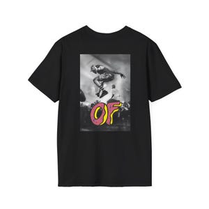 May include: Black t-shirt featuring a graphic of a person mid-air above a crowd. The graphic includes the letters "OF" in pink and yellow. The image is in black and white, with the exception of the letters.