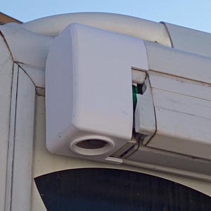 Puede incluir: Un componente de plástico blanco, rectangular, con una abertura circular. Está montado sobre una superficie metálica curvada. La imagen muestra un primer plano del objeto, destacando su forma y textura.