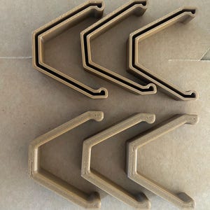 Op de afbeelding: Zes identieke V-vormige clips in een lichtbruine kleur. Elke clip heeft een hoekig ontwerp met een hol midden en kleine, afgeronde haakvormige uiteinden. De clips zijn gerangschikt in twee rijen van drie en hebben een matte afwerking.