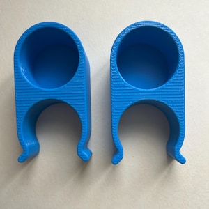 Peut inclure: Deux clips bleu vif, imprimés en 3D, semi-circulaires. Chaque clip possède une ouverture cylindrique en haut et un fond incurvé en forme de griffe. La surface présente une texture striée. Les clips sont conçus pour maintenir ou fixer des objets.