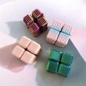 Puede incluir: Vista aérea de cuatro conjuntos de cubos fidget de varios colores. Un conjunto es morado y dorado con una textura espinosa. Otro es rosa y azul con un patrón de panal. El tercero es rosa con círculos concéntricos, y el último es verde y azul con líneas diagonales.