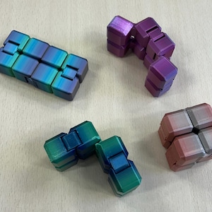 Puede incluir: Varios juguetes fidget de colores están dispuestos sobre una superficie clara. Los juguetes están hechos de cubos conectados en tonos de azul, verde, morado y rosa. Los juguetes están diseñados para ser manipulados y jugados.