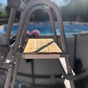 Peut inclure: Une échelle de piscine en métal gris avec une marche en bois. L'échelle est placée devant une piscine hors sol bleue. La marche en bois est faite de planches brun clair.