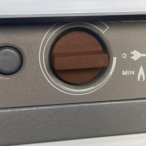 Peut inclure: Gros plan sur un panneau de commande gris d'appareil avec un bouton marron. Le bouton est entouré d'un cercle blanc. Le mot "MIN" est visible, ainsi qu'une icône de flamme et un symbole de prise électrique.