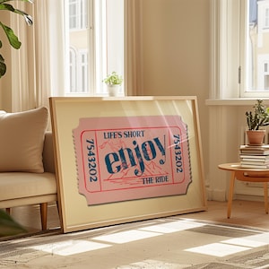 Pode incluir: Impressão emoldurada com um design de bilhete rosa. O texto diz "LIFE'S SHORT ENJOY THE RIDE" em azul e branco. A impressão é exibida em uma sala com luz natural, perto de uma janela.