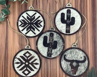 May include: A collection of five round Christmas ornaments with a rustic, southwestern theme. Each ornament features a unique design, including a snowflake, cactus, and bull skull, set against a cowhide-like background with a wooden frame and twine hanger.