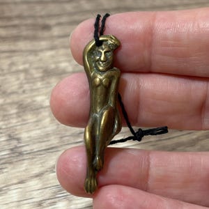May include: A small, antique brass pendant in the shape of a stylized figure, possibly a mermaid or a mythical being. The figure is suspended from a black cord. The pendant has a detailed face and body.