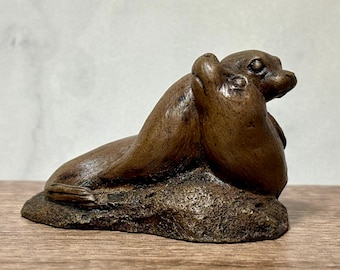 May include: A brown sculpture of two seals resting together on a textured rock base. The seals are positioned side by side with their heads turned slightly, featuring smooth skin textures and detailed facial features in a dark brown finish.