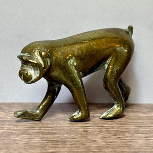 Puede incluir: Una pequeña figura de latón macizo que representa a un mono de pie a cuatro patas. El metal tiene una pátina de color marrón dorado desgastada con una textura superficial visible y reflejos en la espalda, las extremidades y la cara.