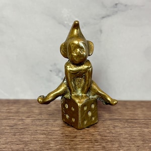 May include: A small, brass-coloured figurine of a seated elf with outstretched legs, perched atop a dice. The elf wears a pointed hat and has large ears. The dice has visible dot markings. The figurine is on a wooden surface.