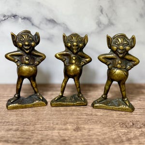 May include: Three antique brass pixie figurines. Each figure has a smiling face, pointed ears, and is posed with hands on hips. The pixies stand on a small base with the word "PIXIE" or "J.C.N.T" engraved.