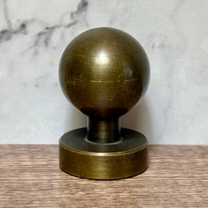 May include: A spherical brass knob with a brushed finish, featuring a round top section connected to a cylindrical neck and a flat, circular base. The metal surface displays subtle horizontal striations and a warm, muted golden-brown tone.