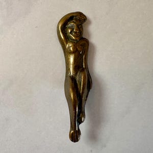 May include: A bronze-colored figurine of a nude person in a relaxed pose, with one arm raised above the head. The figure has a smiling face and is detailed with visible anatomical features. The figurine is likely a decorative item.