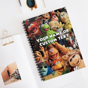 May include: A spiral-bound notebook featuring a collage of Muppet characters. The cover displays Kermit the Frog, Fozzie Bear, Miss Piggy, and others. The text "YOUR NAME OR CUSTOM TEXT" is printed on the cover. The notebook is open on a white surface.