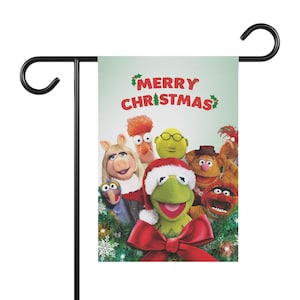 May include: A green and white Christmas garden flag featuring the Muppets. The flag has the words "MERRY CHRISTMAS" in red. Kermit the Frog is wearing a Santa hat and a red bow. The flag is attached to a black metal stand.
