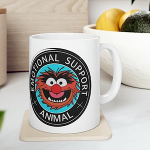 May include: White ceramic mug with a black circle graphic featuring a cartoon red and orange monster with a blue background. The circle graphic has the text "Emotional Support Animal" in black.