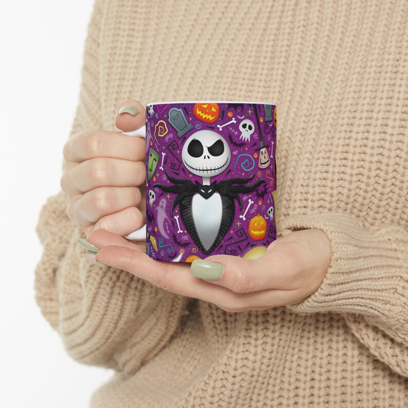 May include: White ceramic mug with a Halloween design. The mug features Jack Skellington, surrounded by pumpkins, ghosts, and other spooky elements on a purple background. The mug is held by a person.