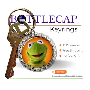 May include: A green frog face on a silver bottle cap keychain. The keychain is attached to a gold key. The text "BOTTLECAP Keyrings" is in the background.
