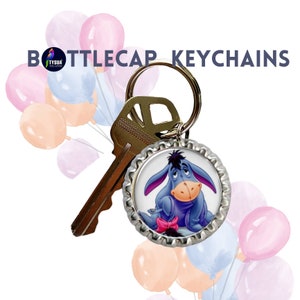 May include: A blue and purple cartoon donkey character on a silver bottle cap keychain. The keychain is attached to a silver key ring. The background is a white surface with pink and blue balloons.