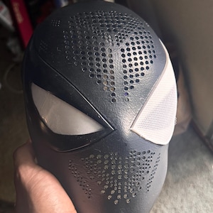 May include: A black full-face mask featuring a textured surface with perforated ventilation holes across the forehead and chin areas. The mask includes two large, angular eye openings covered with a white, fine-mesh material for visibility.
