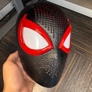 May include: A black full-face mask featuring a textured surface with small circular perforations across the forehead and nose area. The mask includes two large, almond-shaped eye openings outlined in bright red with white mesh lenses.