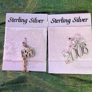 Puede incluir: Dos colgantes de plata de ley en tarjetas blancas. Un colgante dice "GRAD" con una cadena dorada. El otro colgante es el número "2006". Ambas tarjetas dicen "Sterling Silver" y "Tarnish Free".