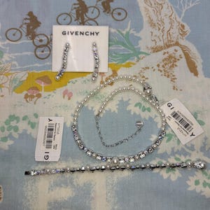 May include: A selection of Givenchy jewellery, including silver earrings, a pearl necklace, and a bracelet. The earrings and bracelet feature clear crystals. The necklace has a mix of pearls and crystals. The Givenchy logo is visible.