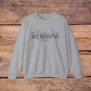 May include: A light gray sweatshirt with the text "Banff National Park CANADA" in dark blue. Delicate purple flower illustrations are above the text. The sweatshirt is made of a soft material.