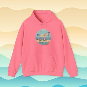 May include: A pink hooded sweatshirt with a front pocket and a drawstring hood. The sweatshirt features a circular graphic of a beach scene with palm trees, beach chairs, and an umbrella. The graphic is in shades of blue, yellow, and tan.