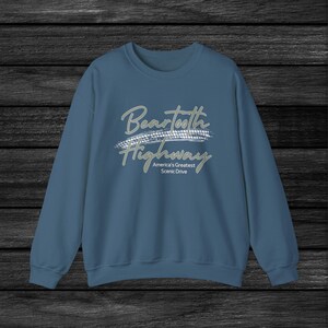 以下が含まれることがあります： 「Beartooth Highway」とタイヤのトレッドデザイン、そして「America's Greatest Scenic Drive」というテキストが書かれた白いグラフィックプリントが施された青いスウェットシャツです。
