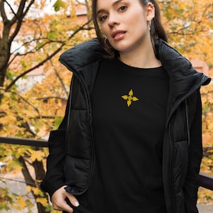 May include: Black long-sleeve shirt with a gold emblem on the chest. The emblem is a four-pointed star design with a floral pattern. The shirt is worn with a black puffer vest. The background features autumn foliage.