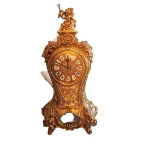 May include: A gold-toned mantle clock with an ornate design and a cherub figure on top. The clock face has Roman numerals and a black hour and minute hand.