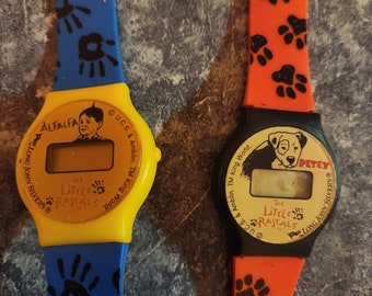 Könnte beinhalten: Zwei bunte Armbanduhren mit schwarzen Digitalanzeigen. Die Uhr links hat ein gelbes Zifferblatt mit den Worten "The Little Rascals" und "ALFALFA" darauf gedruckt. Die Uhr rechts hat ein schwarzes Zifferblatt mit den Worten "The Little Rascals" und "PETEY" darauf gedruckt.