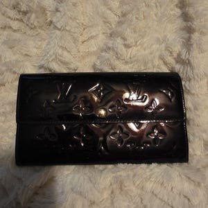 May include: A black patent leather wallet with embossed floral and logo patterns. The wallet has a fold-over flap and a gold-toned snap closure. The surface reflects light, highlighting the embossed design. The wallet is rectangular and appears to be new.
