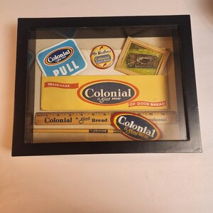 Può includere: Una teca incorniciata di cimeli pubblicitari vintage di Colonial Bread. La teca include un coperchio di scatola di pane Colonial giallo e rosso, un righello con le parole "Colonial is good Bread", una matita con la parola "Colonial" stampata su di essa e un piccolo bottone rotondo con le parole "Mr. Vantness Colonial Is Good Bread".
