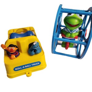 Pode incluir: Um caminhão de brinquedo amarelo com interior azul e uma função de rolar bola azul e amarela. O caminhão tem um personagem da Vila Sésamo, Cookie Monster, no banco do motorista e outro personagem da Vila Sésamo, Elmo, no banco do passageiro. O caminhão tem as palavras "Roll-A-Ball Truck" impressas na frente. Um brinquedo azul com um personagem de sapo verde dentro. O sapo está vestindo uma camisa listrada azul e branca e está andando de triciclo vermelho.