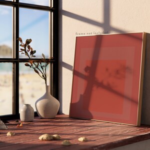 Op de afbeelding: Een ingelijst abstract schilderij met een rood, oranje en roze kleurenschema. Het schilderij leunt tegen een witte muur, met een raam en een strandsc&egrave;ne op de achtergrond. Er staan ook twee kleine vazen en wat schelpen op een houten oppervlak.