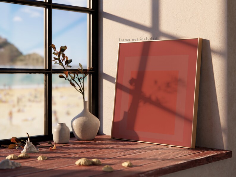 Op de afbeelding: Een ingelijst abstract schilderij met een rood, oranje en roze kleurenschema. Het schilderij leunt tegen een witte muur, met een raam en een strandsc&egrave;ne op de achtergrond. Er staan ook twee kleine vazen en wat schelpen op een houten oppervlak.