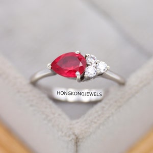 Könnte beinhalten: Ein silberner Ring mit einem birnenförmigen, leuchtend roten Edelstein als Mittelpunkt, ergänzt durch drei kleinere, klare Edelsteine, die sich daneben befinden. Der Ring hat ein schlankes, poliertes Band. Der Text "HONGKONGJEWELS" ist sichtbar.