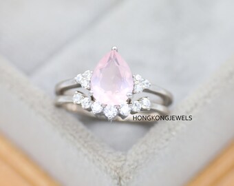 May include: A silver ring set featuring a pear-shaped, light pink gemstone as the centerpiece, surrounded by smaller, clear stones. The ring is accompanied by a matching band. The text "HONGKONGJEWELS" is visible.