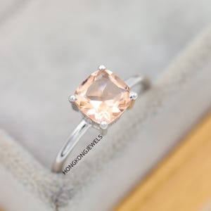 Puede incluir: Un anillo de plata con una gema de color melocotón, talla cojín. La gema está engastada con cuatro garras. El anillo tiene una banda delgada. El texto "HONGKONGJEWELS" es visible en la banda del anillo.