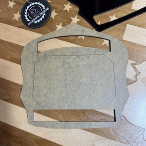 Puede incluir: Un recorte de madera de un escudo de bomberos con un espacio en blanco para la personalización. El escudo es marrón y tiene un recorte para un nombre o un logotipo.