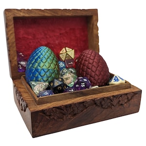 May include: A wooden box with a red velvet interior, filled with colorful dice and two dragon egg-shaped objects. One egg is blue and green, the other is red.