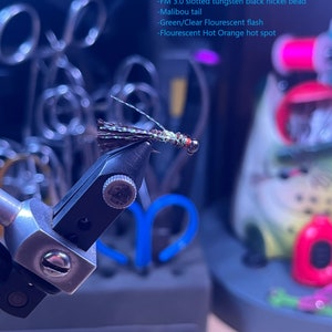 May include: A black nickel micro jig streamer fly fishing lure with a green and clear fluorescent flash and a hot orange spot. The fly has a black nickel bead and a malibou tail.