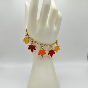 May include: Gold-toned chain bracelet with six enamel maple leaf charms in shades of red, orange, and yellow. The bracelet is displayed on a white mannequin hand against a white background. A fall-themed jewelry item.