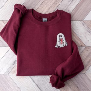 May include: Burgundy-colored sweatshirt with a white embroidered ghost design holding a can of soda. The ghost has two oval eyes and a rectangular mouth. The sweatshirt has long sleeves and a crew neck.