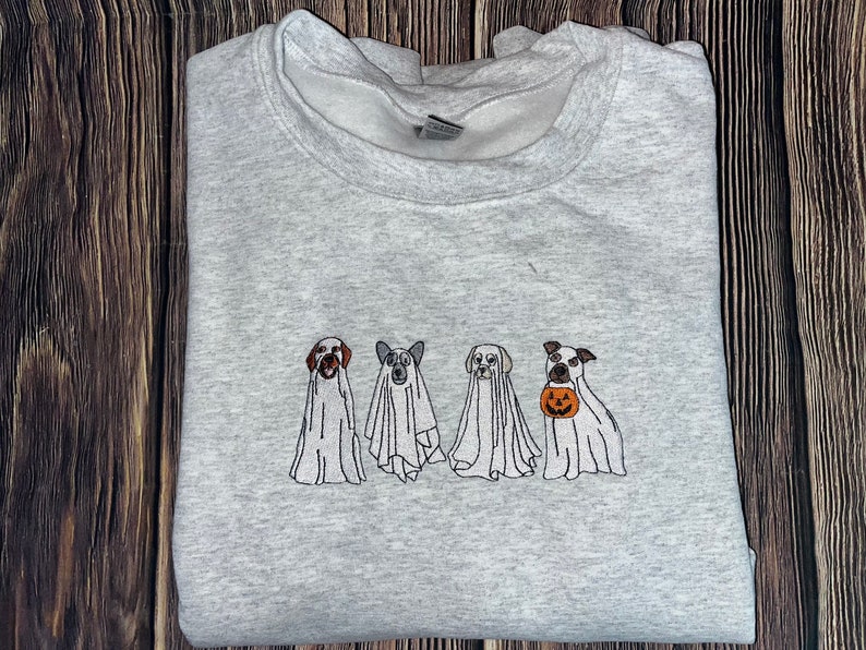 以下が含まれることがあります： ハロウィン用のゴーストに扮した4匹の犬が刺繍されたグレーのトレーナー。3匹の犬は白いゴーストの衣装を着ており、1匹の犬は小さなオレンジ色のカボチャを持っています。